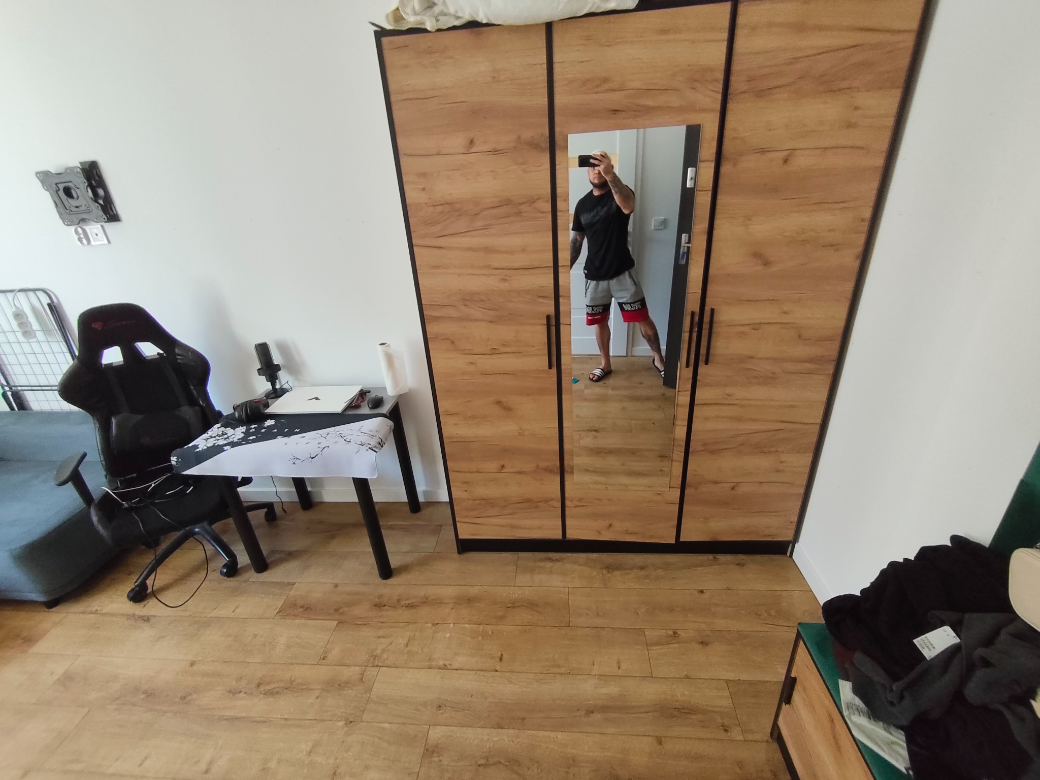 Pokój z drewnianą podłogą i szafą z lustrem, w którym odbija się mężczyzna robiący zdjęcie telefonem. Widoczne biurko z krzesłem gamingowym i uchwyt na telewizor na ścianie.