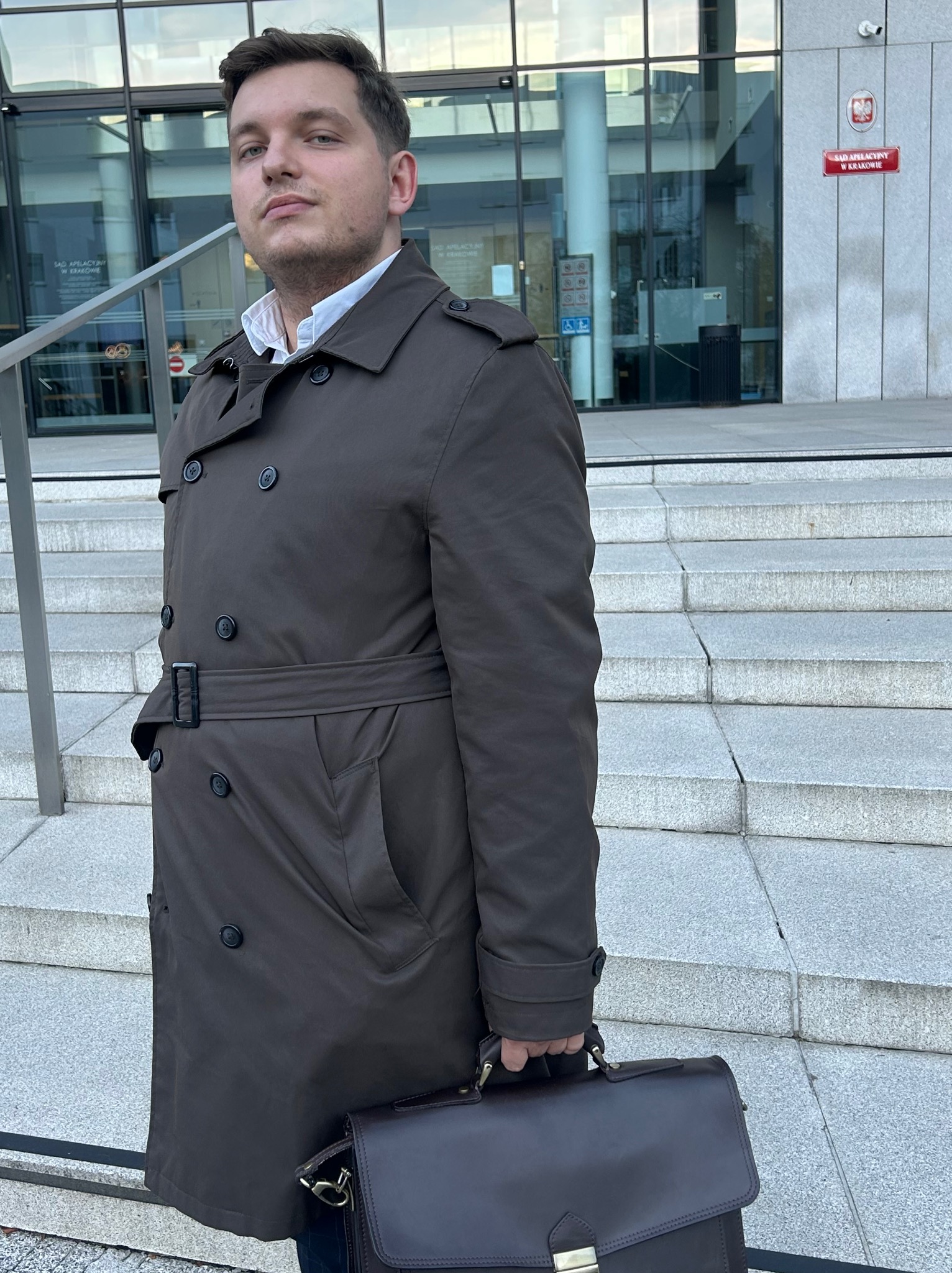 Młody mężczyzna w płaszczu z aktówką przed Sądem Apelacyjnym w Krakowie. Elegancki prawnik w drodze na rozprawę. Profesjonalizm i powaga.