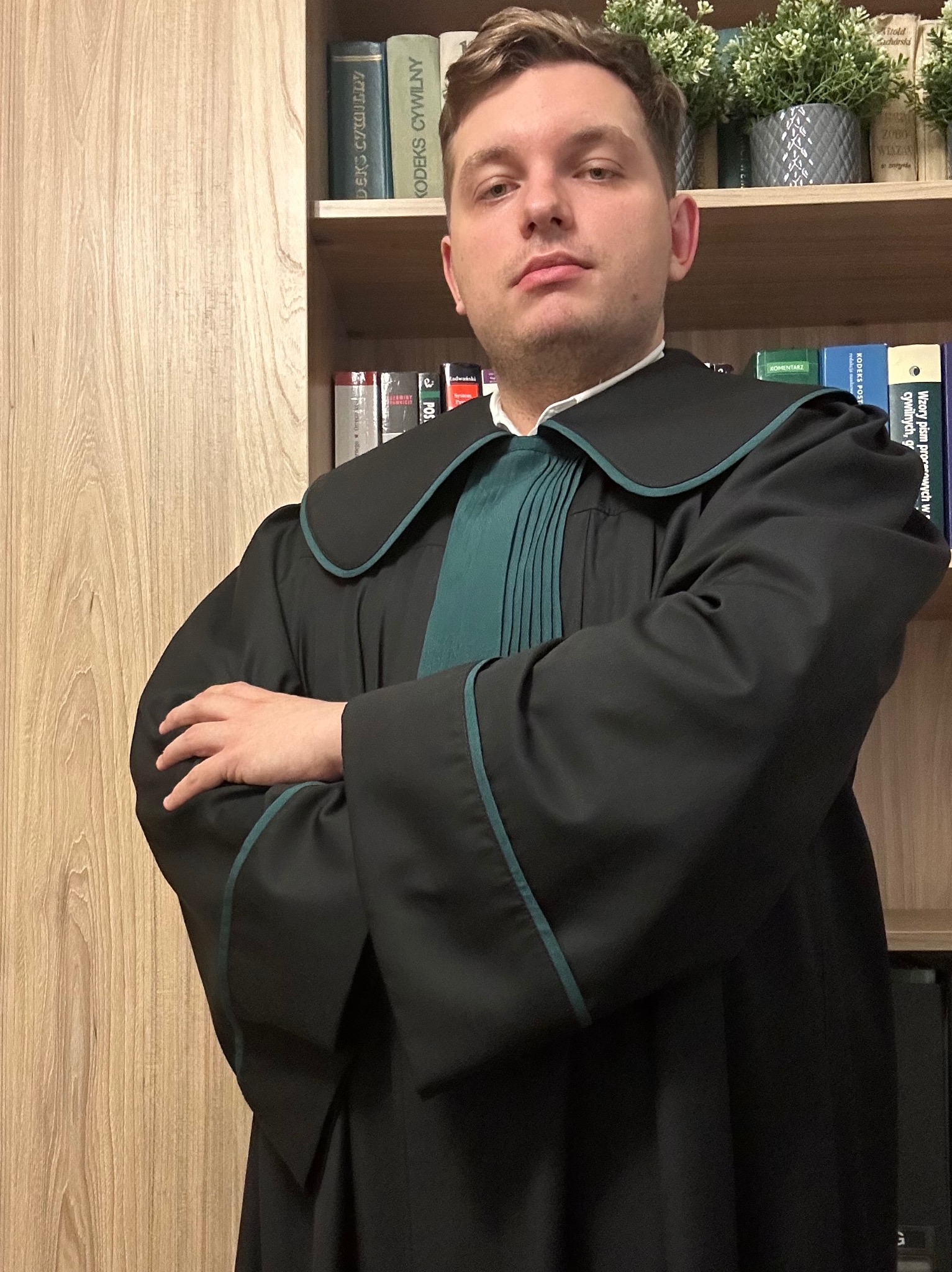 Portret młodego adwokata w todze na tle regału z książkami prawniczymi i dekoracjami roślinnymi. Ujęcie od pasa w górę, skrzyżowane ręce, poważny wyraz twarzy.