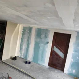 Wnętrze w trakcie prac wykończeniowych: ściany z płyt gipsowych, szpachlowane łączenia, widoczna drewniana stolarka drzwiowa z szybą i sufit w trakcie obróbki.