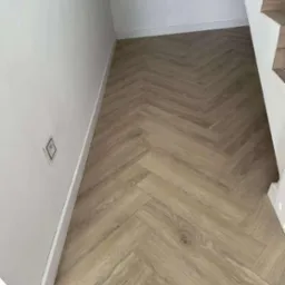 Jasne panele ułożone w jodełkę francuską, przy białej ścianie z gniazdkiem i fragmentem schodów. Widok na podłogę po montażu, z bliska.