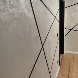 USŁUGI OGÓLNOBUDOWLANE Mateusz Grodkowski - Ściana z dekoracyjną fakturą i czarnymi, geometrycznymi liniami, widok z boku na czarną ościeżnicę drzwiową i fragment podłogi z paneli.