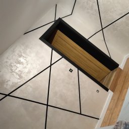 USŁUGI OGÓLNOBUDOWLANE Mateusz Grodkowski - Dekoracyjna ściana z efektem betonu i czarnymi listwami, oświetlenie sufitowe, widok na drewniane drzwi w czarnej ościeżnicy, fragment blatu z akcesoriami malarskimi.