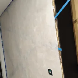 Ściana po szpachlowaniu z widocznymi śladami szpachli, zabezpieczona niebieską taśmą malarską przy suficie i ościeżnicy. Widoczne gniazdka elektryczne.