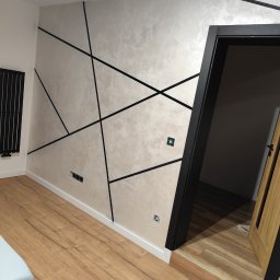 USŁUGI OGÓLNOBUDOWLANE Mateusz Grodkowski - Ściana z dekoracyjną, srebrzystą farbą i geometrycznym wzorem z czarnych listew. Podłoga z paneli drewnopodobnych, czarna ościeżnica drzwiowa.