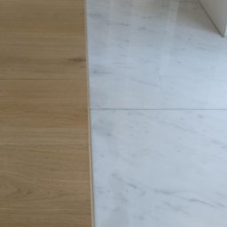 USŁUGI OGÓLNOBUDOWLANE Mateusz Grodkowski - Połączenie jasnych paneli podłogowych i białych płytek marmurowych. Widok na podłogę wzdłuż okna. Kontrast materiałów i kolorów, nowoczesny design.