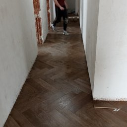 USŁUGI OGÓLNOBUDOWLANE Mateusz Grodkowski - Ułożony parkiet w jodełkę w korytarzu, widoczny pracownik w tle, ściany w trakcie wykończenia.
