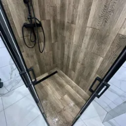 Nowoczesna kabina prysznicowa z drewnopodobnymi płytkami na ścianach i podłodze, czarna armatura i odpływ liniowy, białe płytki poza kabiną.