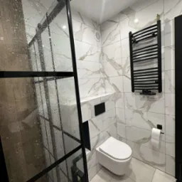 Nowoczesna łazienka z kabiną prysznicową typu walk-in z czarnymi profilami, marmurowe płytki na ścianach, podwieszana toaleta i grzejnik drabinkowy.