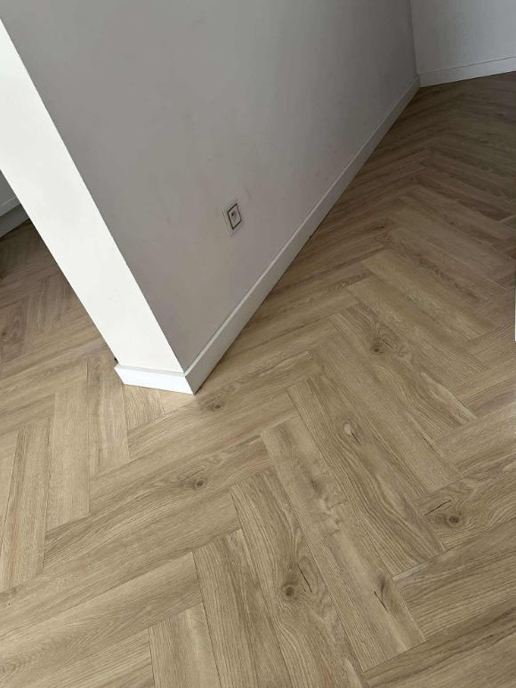 Jasne panele ułożone we wzór jodełki francuskiej, przy białej ścianie z listwą przypodłogową i gniazdkiem. Widok z góry, neutralne oświetlenie.