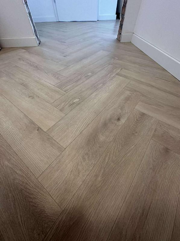 Ułożone panele podłogowe w jodełkę, jasne drewno, białe listwy przypodłogowe, widoczne fragmenty ścian i drzwi. Minimalistyczny kadr na podłogę.