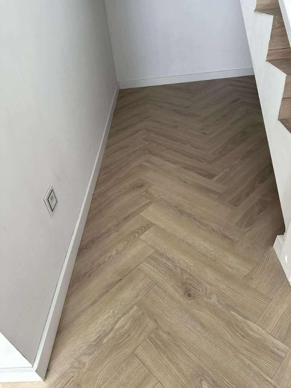 Jasne panele ułożone w jodełkę francuską, przy białej ścianie z gniazdkiem i fragmentem schodów. Widok na podłogę po montażu, z bliska.