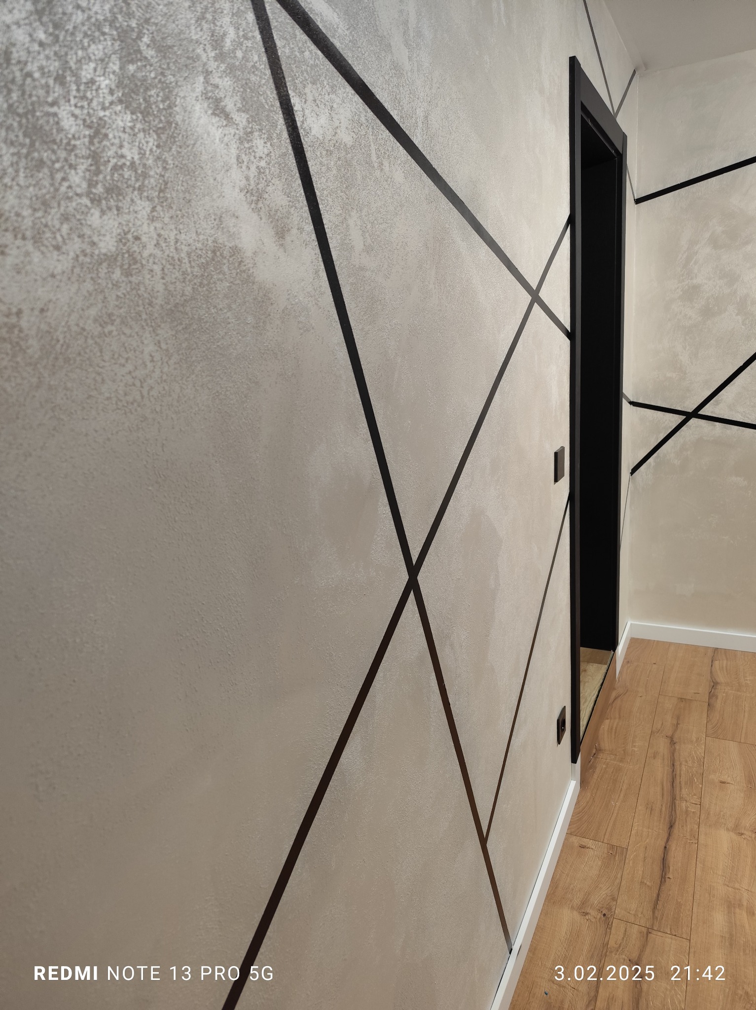 Ściana z dekoracyjną fakturą i czarnymi, geometrycznymi liniami, widok z boku na czarną ościeżnicę drzwiową i fragment podłogi z paneli.