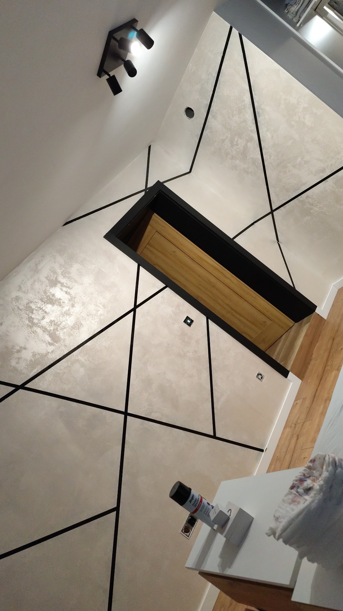 Dekoracyjna ściana z efektem betonu i czarnymi listwami, oświetlenie sufitowe, widok na drewniane drzwi w czarnej ościeżnicy, fragment blatu z akcesoriami malarskimi.