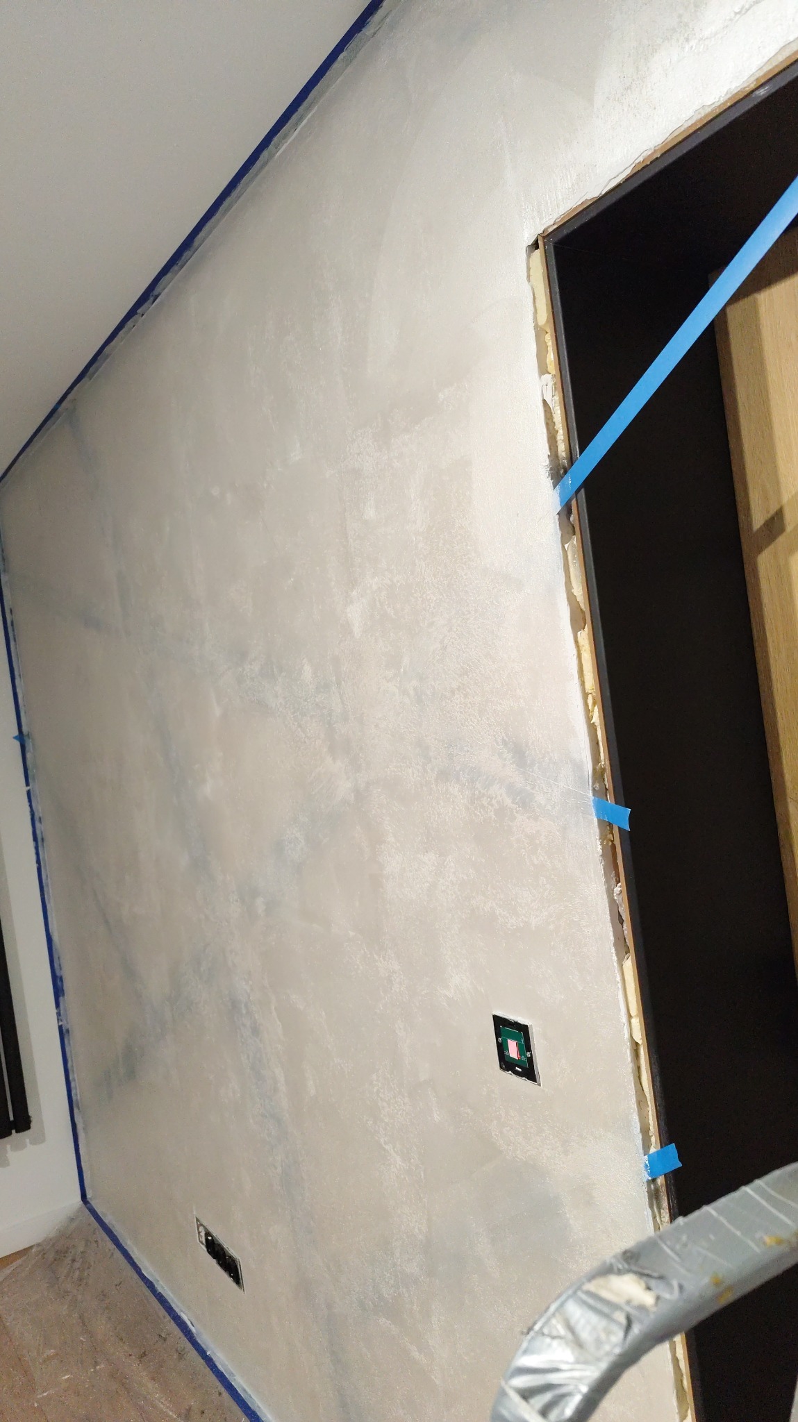 Ściana po szpachlowaniu z widocznymi śladami szpachli, zabezpieczona niebieską taśmą malarską przy suficie i ościeżnicy. Widoczne gniazdka elektryczne.