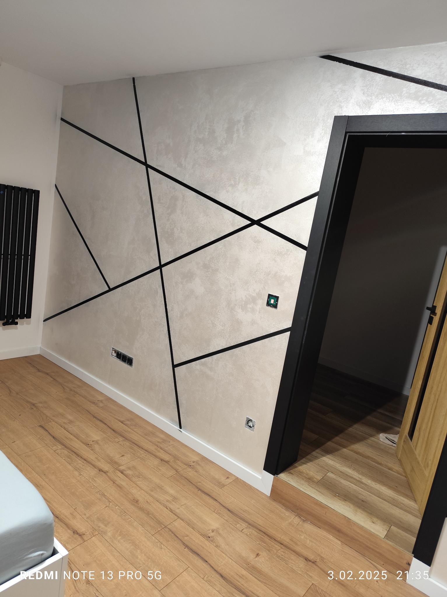 Ściana z dekoracyjną, srebrzystą farbą i geometrycznym wzorem z czarnych listew. Podłoga z paneli drewnopodobnych, czarna ościeżnica drzwiowa.