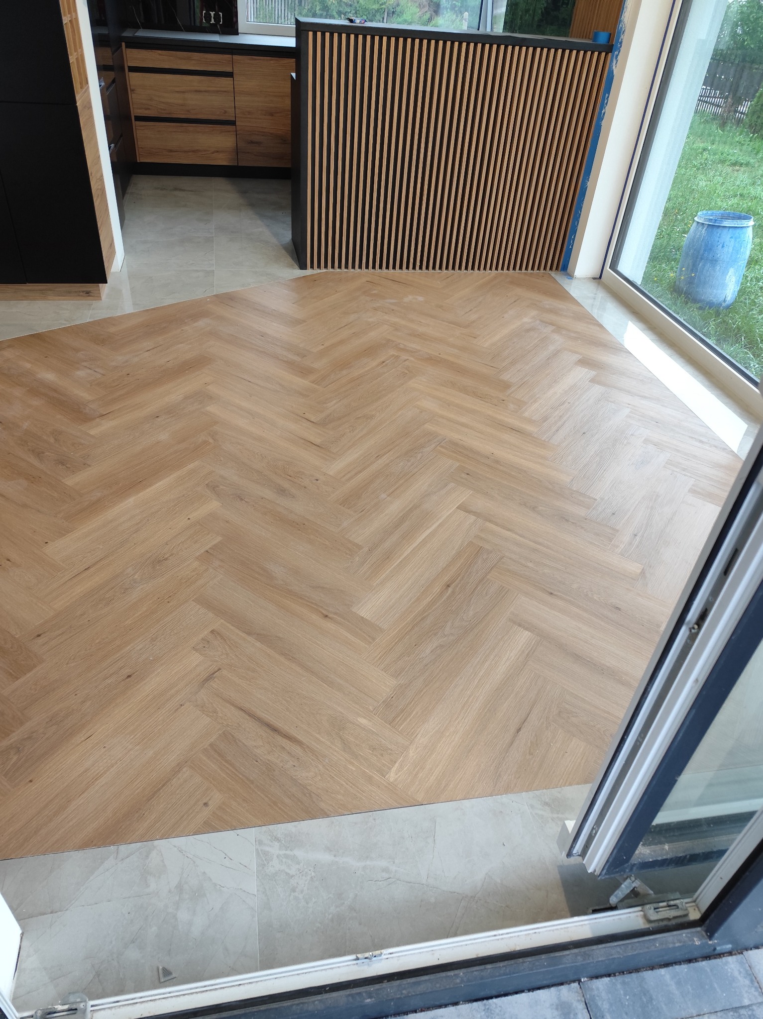 Ułożony parkiet w jodełkę w jasnym odcieniu drewna, z widocznymi fragmentami kuchni i dużym oknem. Nowoczesne wykończenie podłogi.