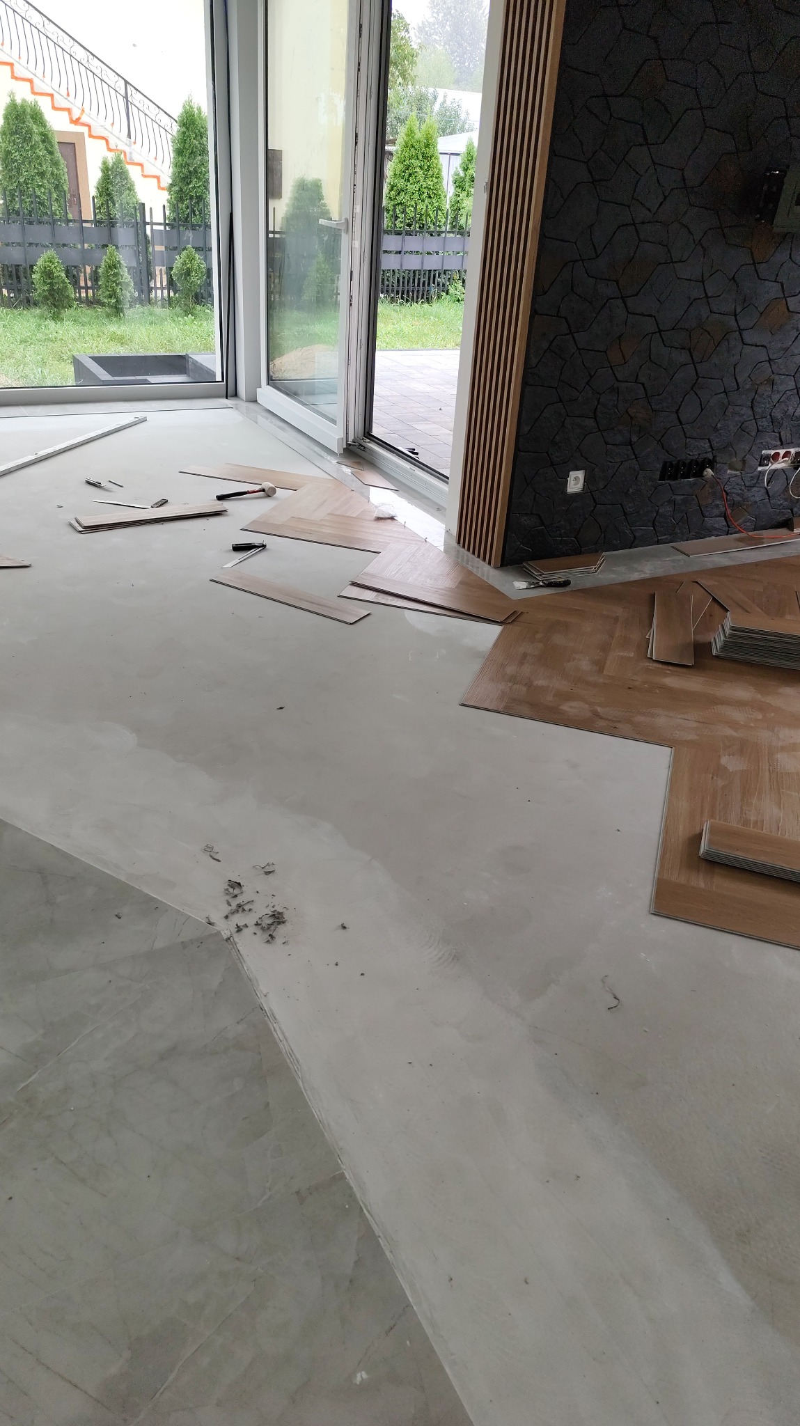 Układanie paneli podłogowych w jodełkę w jasnym wnętrzu z dużym oknem. Widoczne narzędzia i fragment dekoracyjnej ściany z drewnianymi lamelami.