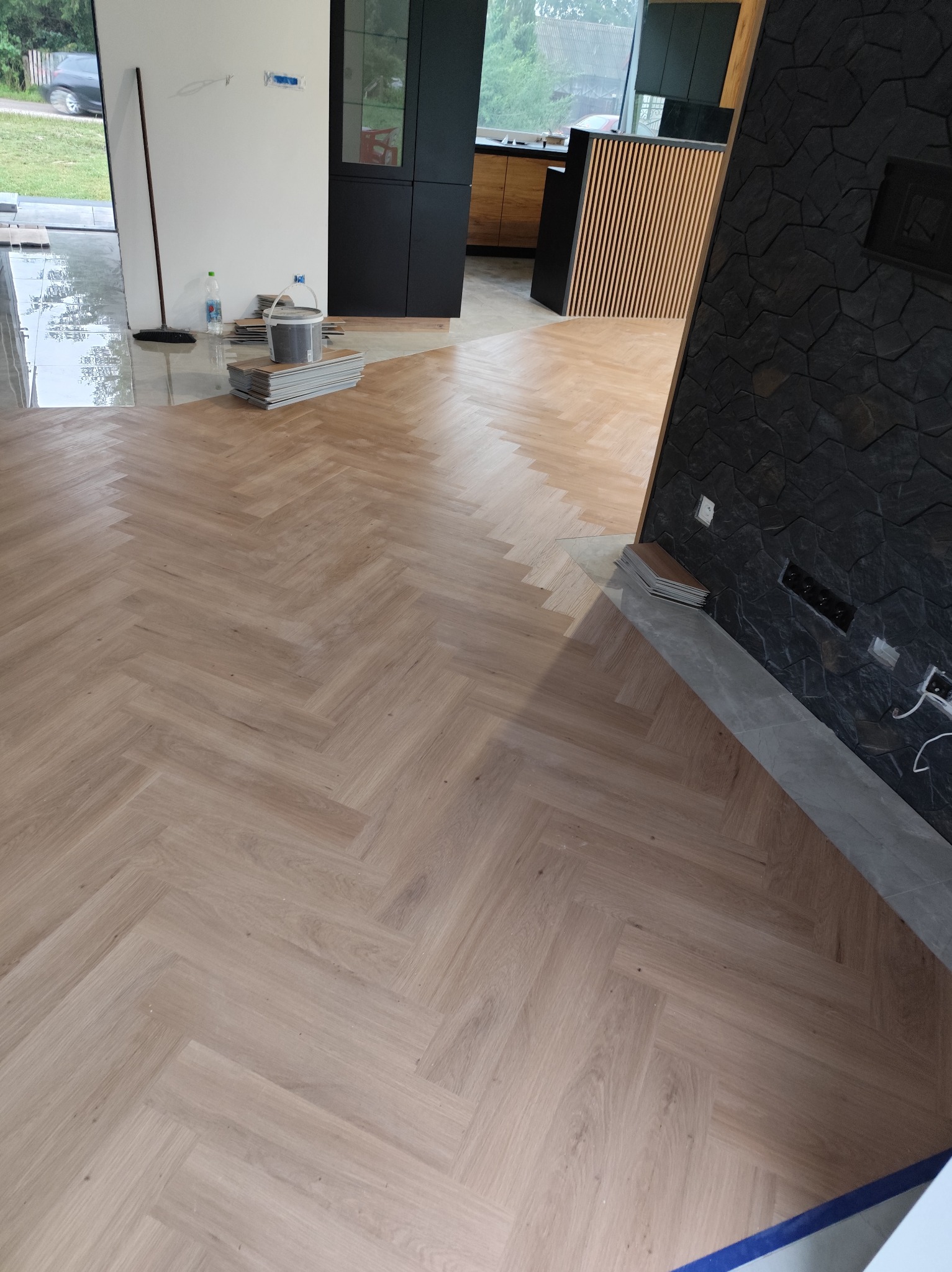 Jasne panele ułożone w jodełkę na podłodze w trakcie prac wykończeniowych. Widoczne narzędzia, resztki paneli i fragment ściany z dekoracyjną okładziną.