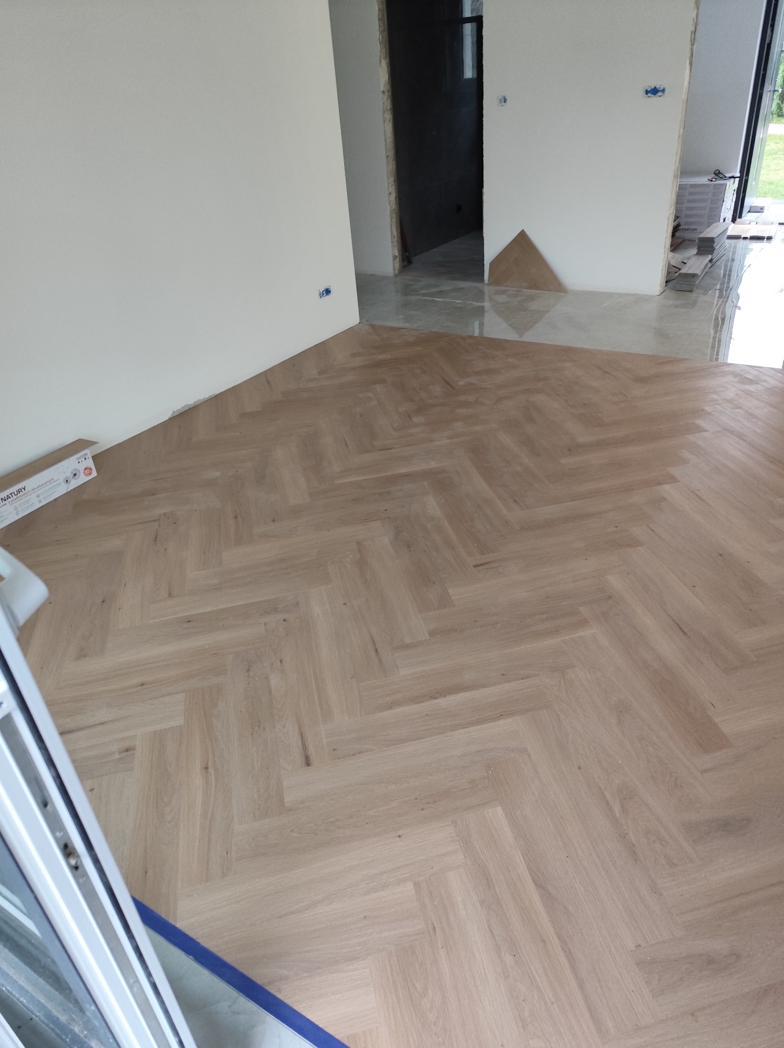 Jasne panele ułożone we wzór jodełki francuskiej w nowym wnętrzu. Widoczne fragmenty ścian i drzwi. Paczka paneli NATURE leży przy ścianie.