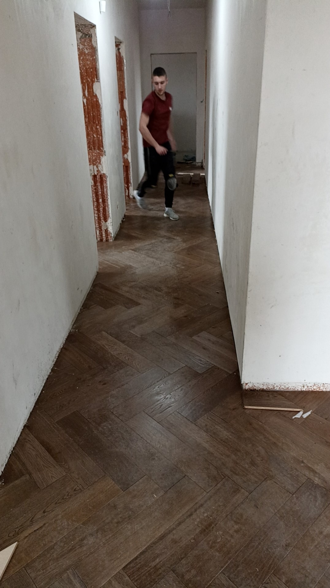 Ułożony parkiet w jodełkę w korytarzu, widoczny pracownik w tle, ściany w trakcie wykończenia.