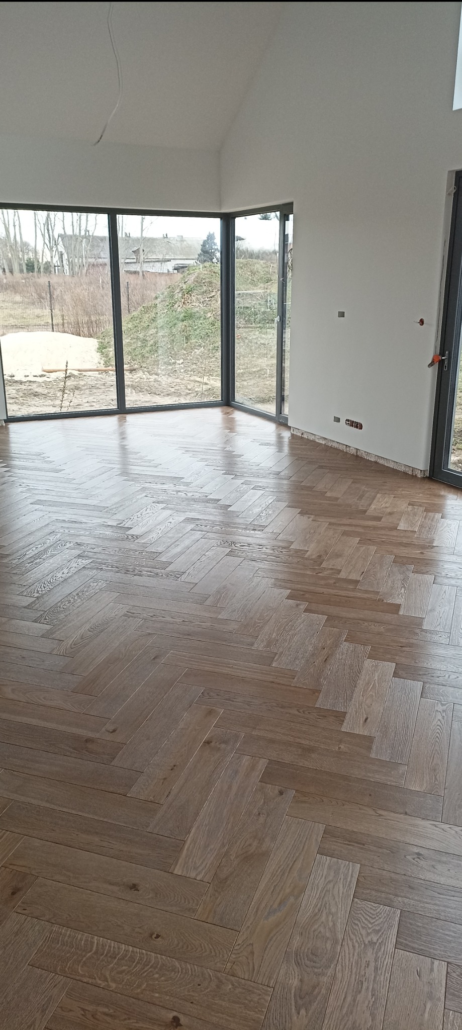 Salon z parkietem ułożonym w jodełkę. Duże okna z widokiem na zewnątrz. Ściany pomalowane na biało. Widoczne gniazdka elektryczne.