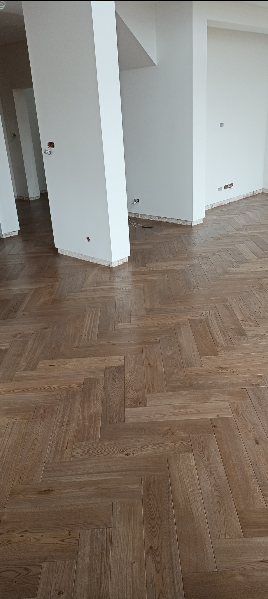 Ukończona podłoga z parkietu ułożonego w jodełkę, w jasnym, przestronnym wnętrzu z białymi ścianami i filarami. Widoczne niedokończone gniazdka elektryczne.