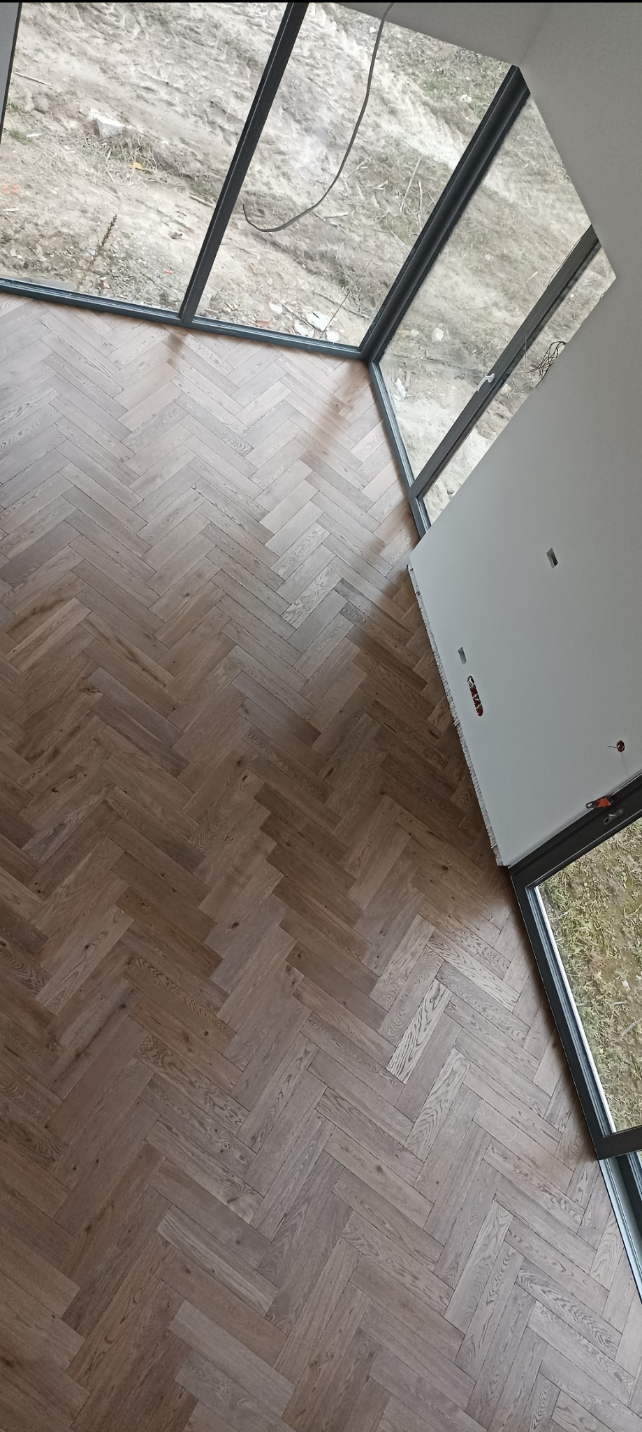 Ułożony parkiet w jodełkę francuską, widok z góry, duże okna z czarnymi ramami, ściana z widocznymi puszkami elektrycznymi. Jasne drewno, surowy krajobraz za oknem.