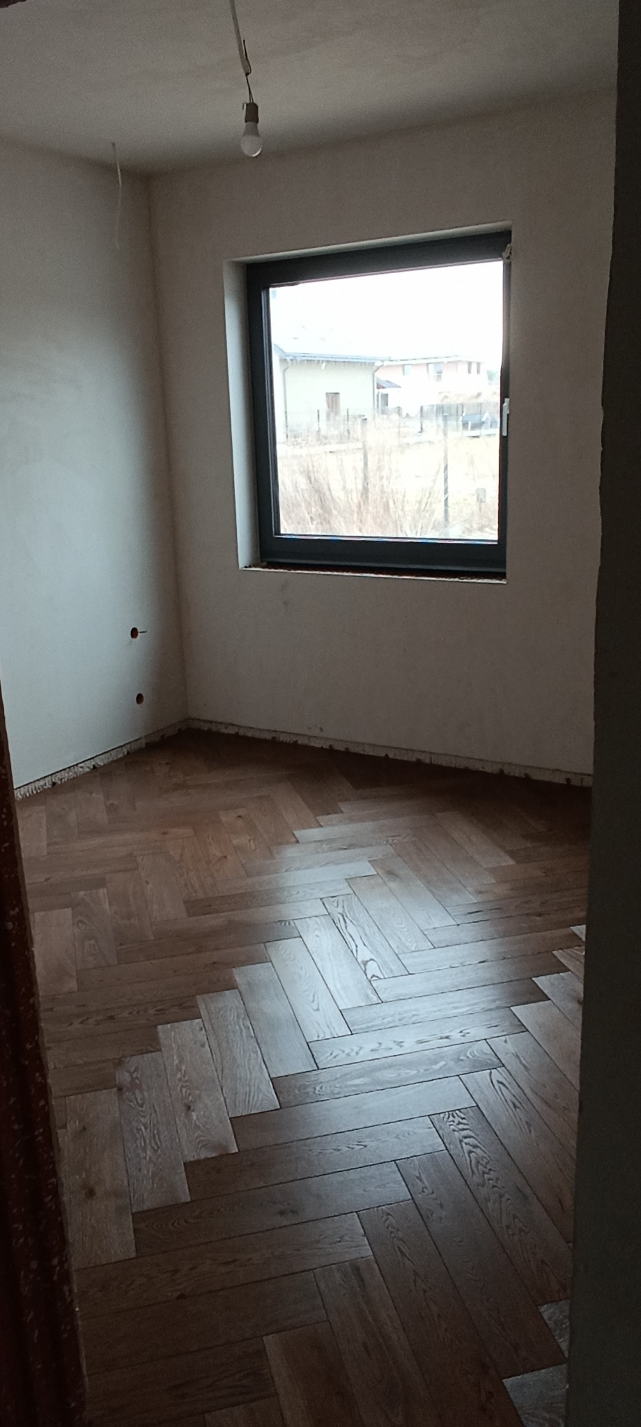 Pokój z ułożonym parkietem w jodełkę, widok na okno z czarną ramą. Ściany przygotowane do malowania, sufit z wiszącą żarówką. Widoczne niedociągnięcia wykończeniowe.