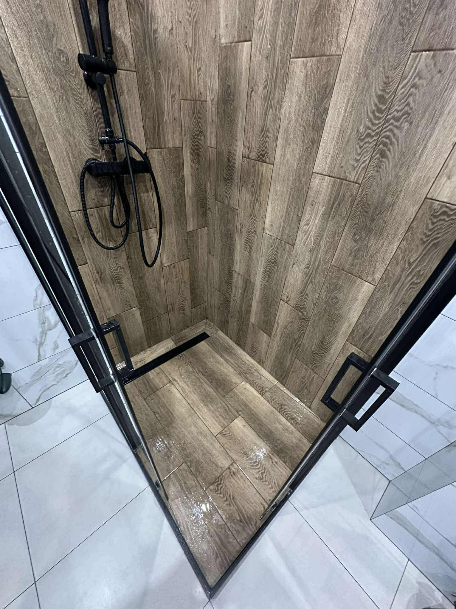 Nowoczesna kabina prysznicowa z drewnopodobnymi płytkami na ścianach i podłodze, czarna armatura i odpływ liniowy, białe płytki poza kabiną.