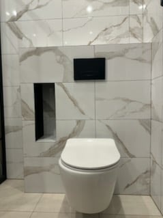 Biała toaleta wisząca na tle marmurowych płytek z czarnym przyciskiem spłukującym i wnęką. Minimalistyczny design łazienki po remoncie.