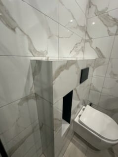 Nowoczesna toaleta podwieszana, obudowana płytami imitującymi marmur, z czarnym przyciskiem spłukującym. Minimalistyczny design, eleganckie wykończenie.