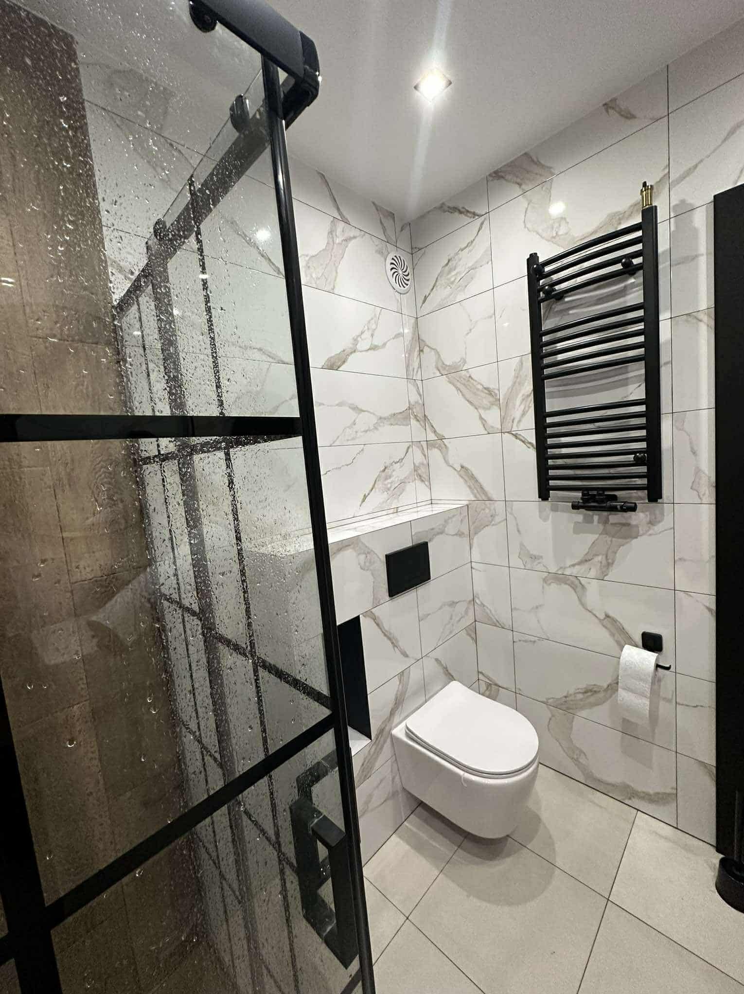 Nowoczesna łazienka z kabiną prysznicową typu walk-in z czarnymi profilami, marmurowe płytki na ścianach, podwieszana toaleta i grzejnik drabinkowy.