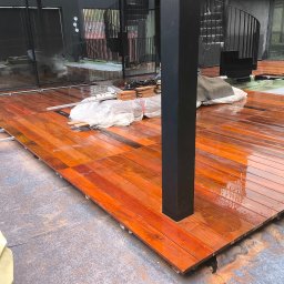 Softwood - Łukasz Wiśniowski - Montaż drewnianego tarasu z egzotycznego drewna. Widoczne deski tarasowe, legary i elementy mocujące. W tle spiralne schody i przeszklenia.