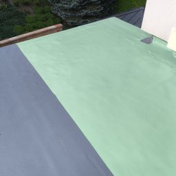 Softwood - Łukasz Wiśniowski - Wykończony dach płaski z hydroizolacją w kolorze szaro-zielonym, widok z góry, z fragmentem zieleni w tle. Detal pokrycia dachowego.