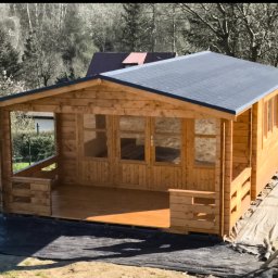 Softwood - Łukasz Wiśniowski - Drewniany domek letniskowy z szarym dachem, widoczny z zewnątrz, z elementami balustrady i przeszklonymi drzwiami, na tle drzew i trawy. Widoczny niebieski pojemnik.