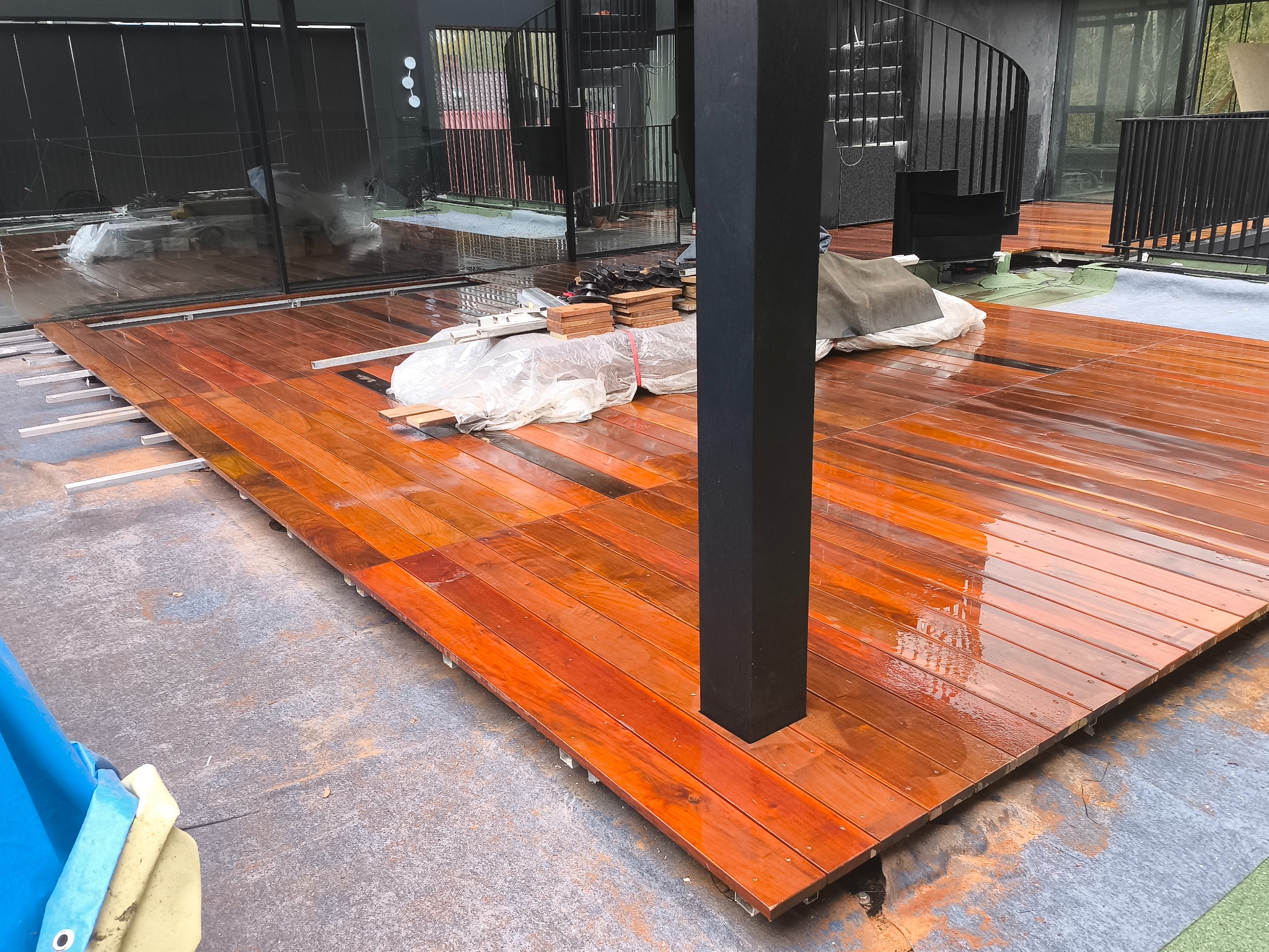 Montaż drewnianego tarasu z egzotycznego drewna. Widoczne deski tarasowe, legary i elementy mocujące. W tle spiralne schody i przeszklenia.