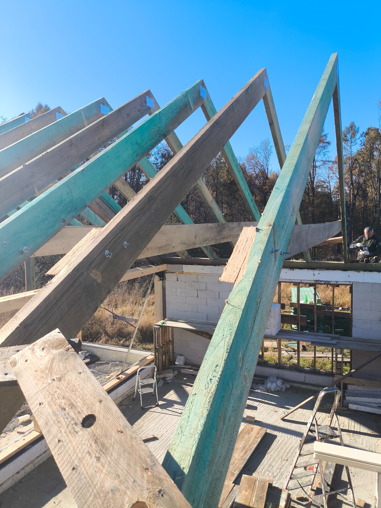 Konstrukcja dachu z drewnianych belek, częściowo pomalowanych na zielono, na tle murowanego budynku w trakcie budowy i błękitnego nieba. Widoczne narzędzia i elementy konstrukcyjne.