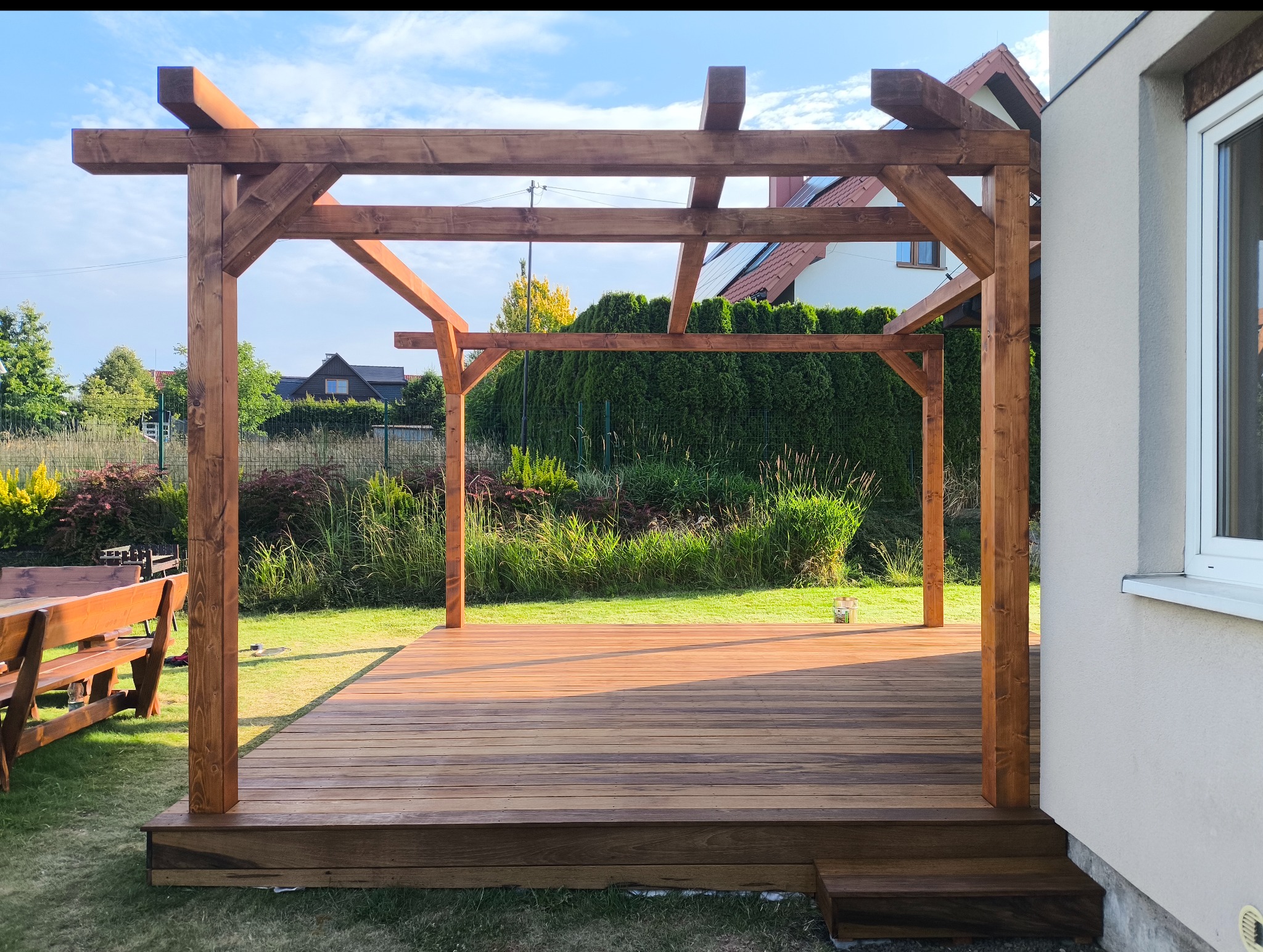 Drewniana pergola z tarasem na trawniku, widok z niskiej perspektywy, z elementami domu w tle. Konstrukcja z belek w ciepłym odcieniu brązu, naturalne otoczenie.