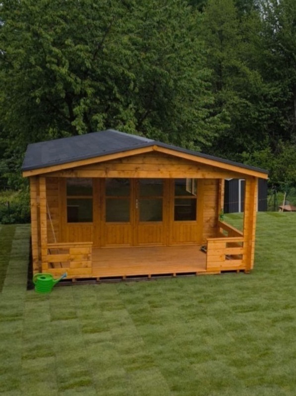 Drewniany domek letniskowy z tarasem w Dąbrówce, otoczony zielonym trawnikiem i drzewami. Widoczny zielony konewka. Konstrukcja z przeszklonymi drzwiami.