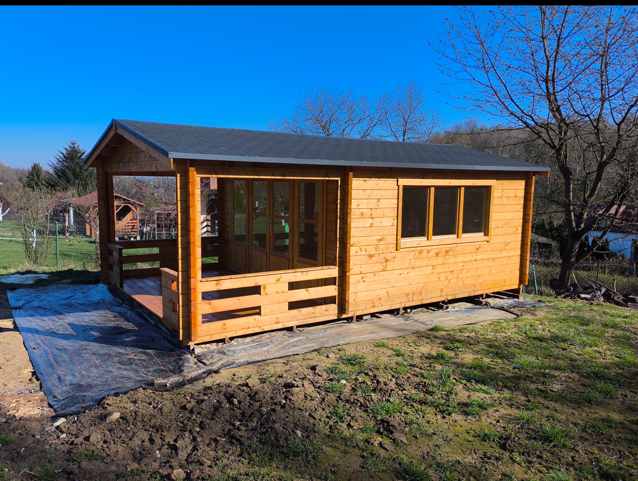 Drewniany domek letniskowy z gontowym dachem w ogrodzie, częściowo na podkładzie z folii. Widoczne okna, drzwi i taras. Błękitne niebo w tle.