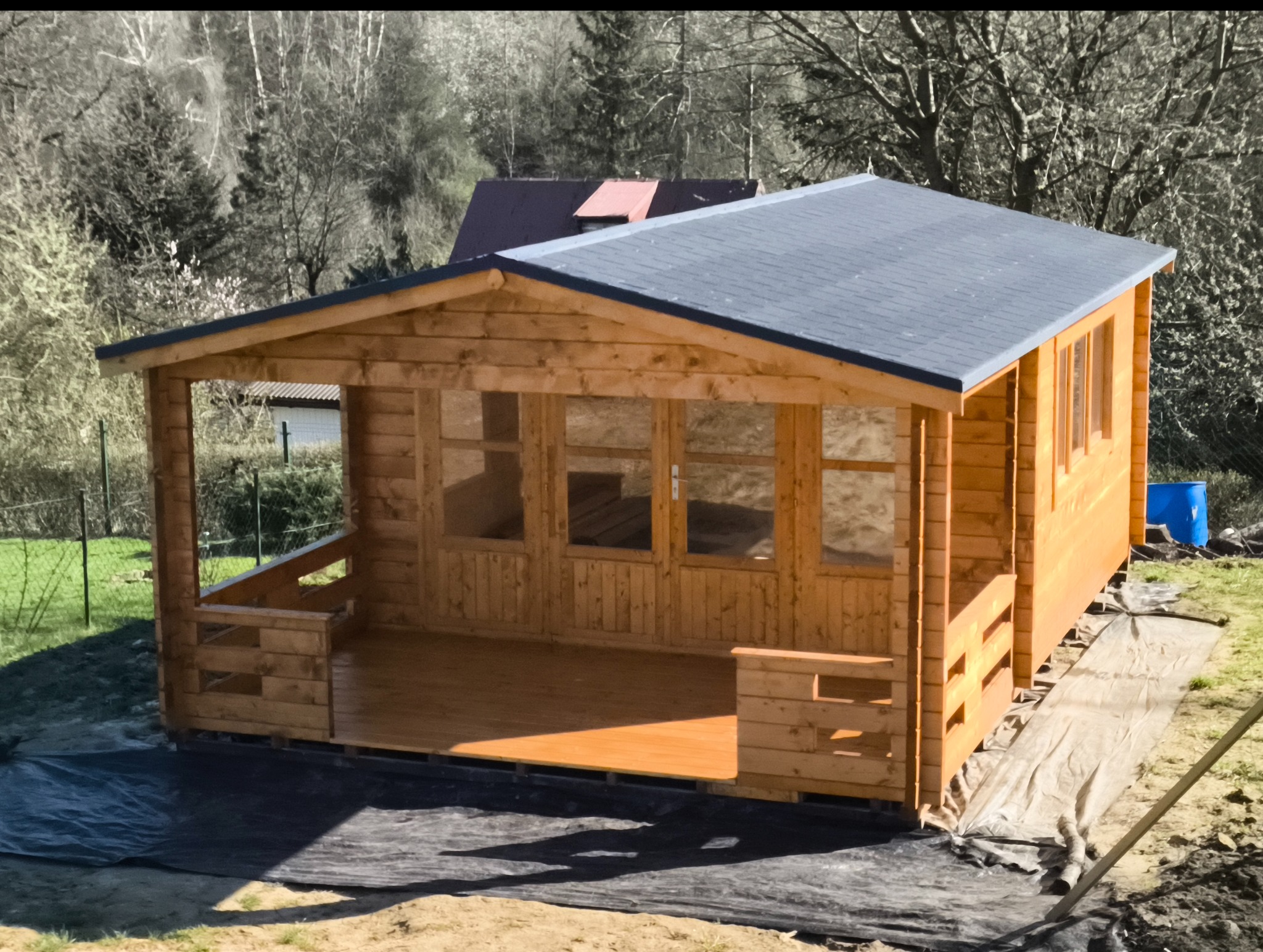 Drewniany domek letniskowy z szarym dachem, widoczny z zewnątrz, z elementami balustrady i przeszklonymi drzwiami, na tle drzew i trawy. Widoczny niebieski pojemnik.