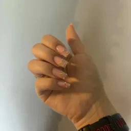 Dłoń z kwadratowymi paznokciami w pastelowych kolorach i z błyszczącymi zdobieniami na tle jasnej ściany. Widoczny zegarek na nadgarstku.