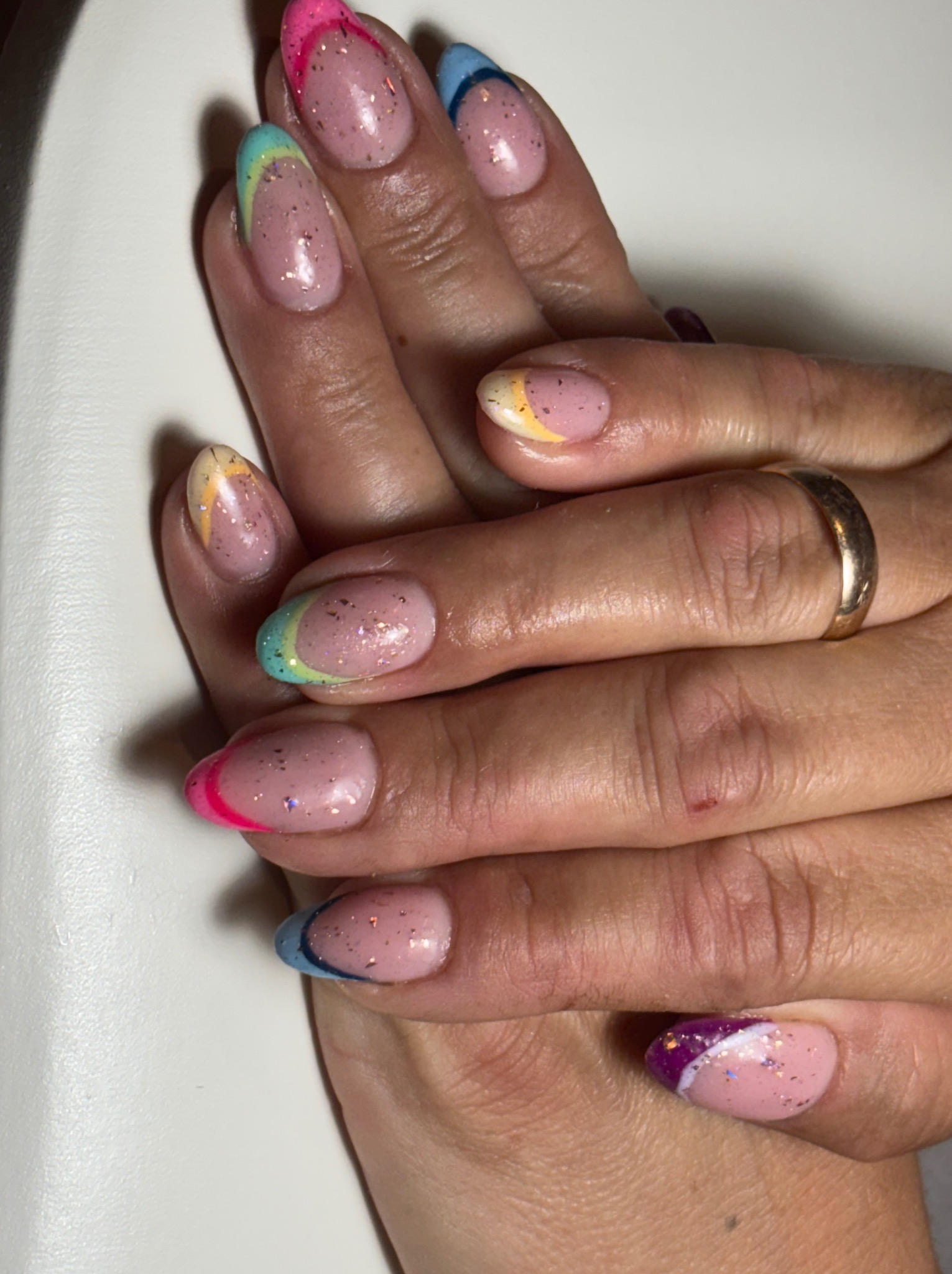 Dłonie z manicure hybrydowym: różowa baza, brokat, kolorowe końcówki french (róż, niebieski, żółty, zielony, fioletowy). Na palcu serdecznym złoty pierścionek.