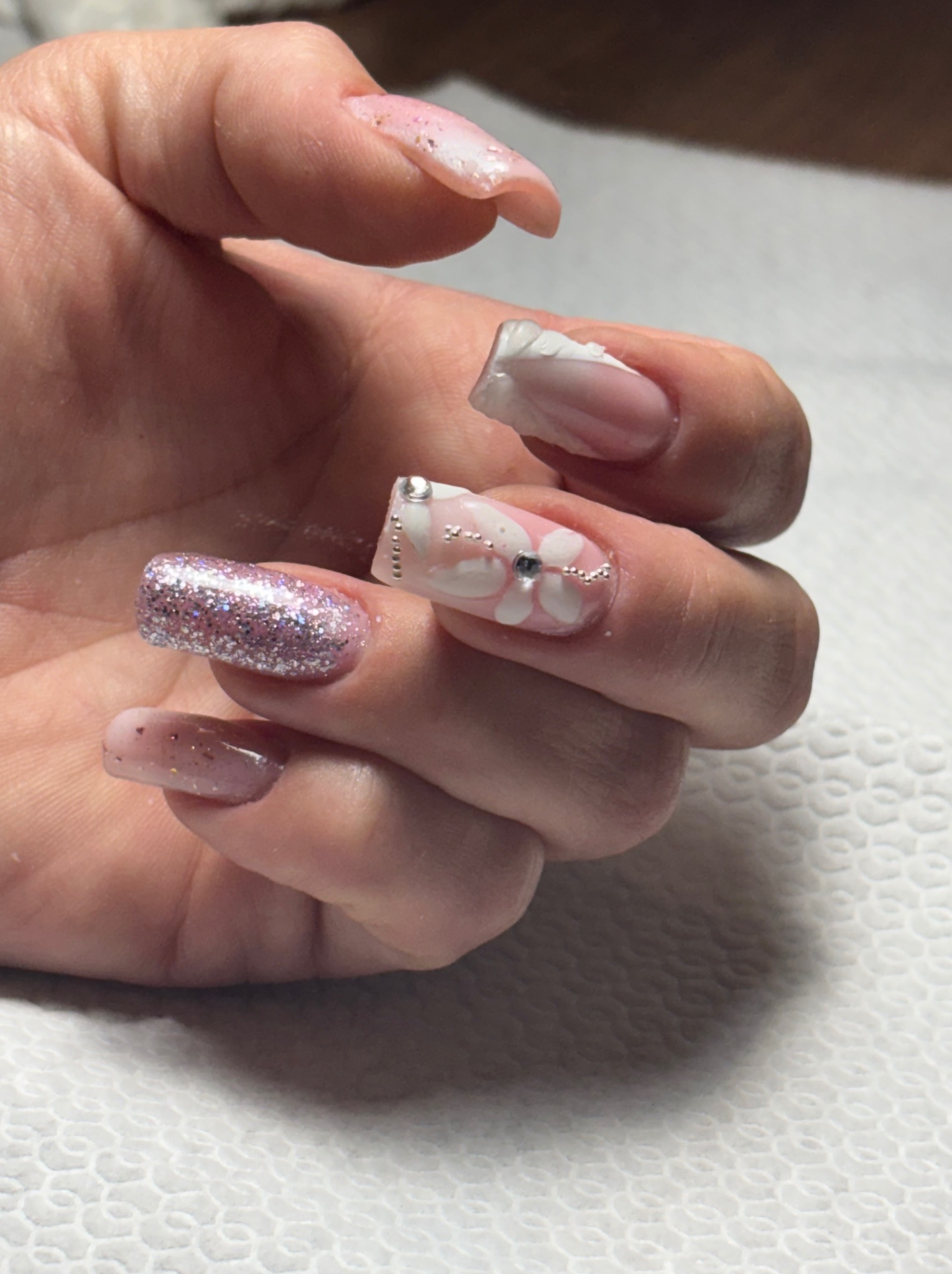 Dłoń z pomalowanymi paznokciami w odcieniach różu i bieli, z brokatem, zdobieniami kwiatowymi i srebrnymi koralikami. Efekt po zabiegu upiększającym.