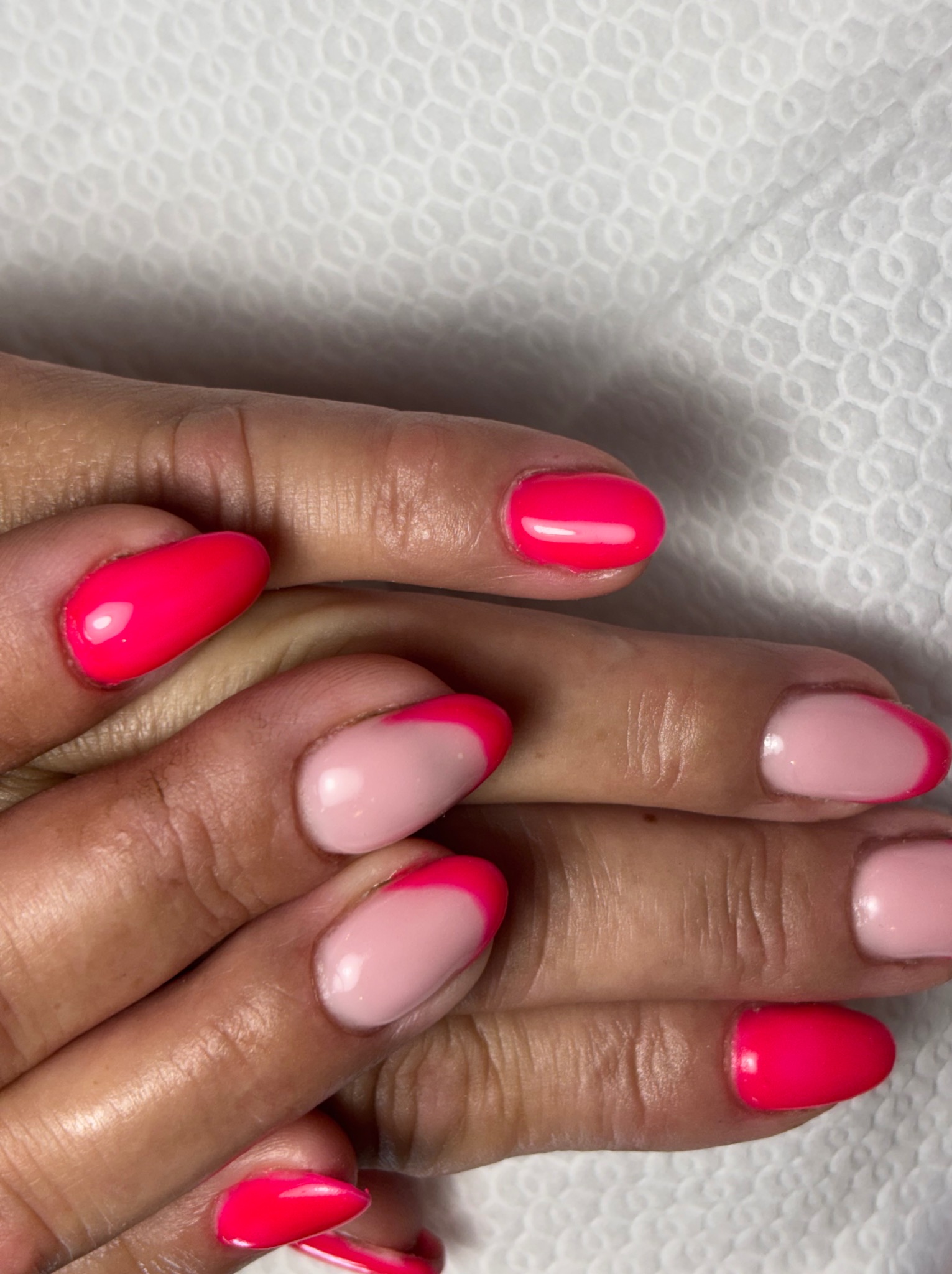 Dłonie z pomalowanymi paznokciami: neonowy róż na całości i delikatny róż z neonowym french manicure na tle białej, teksturowanej podkładki.