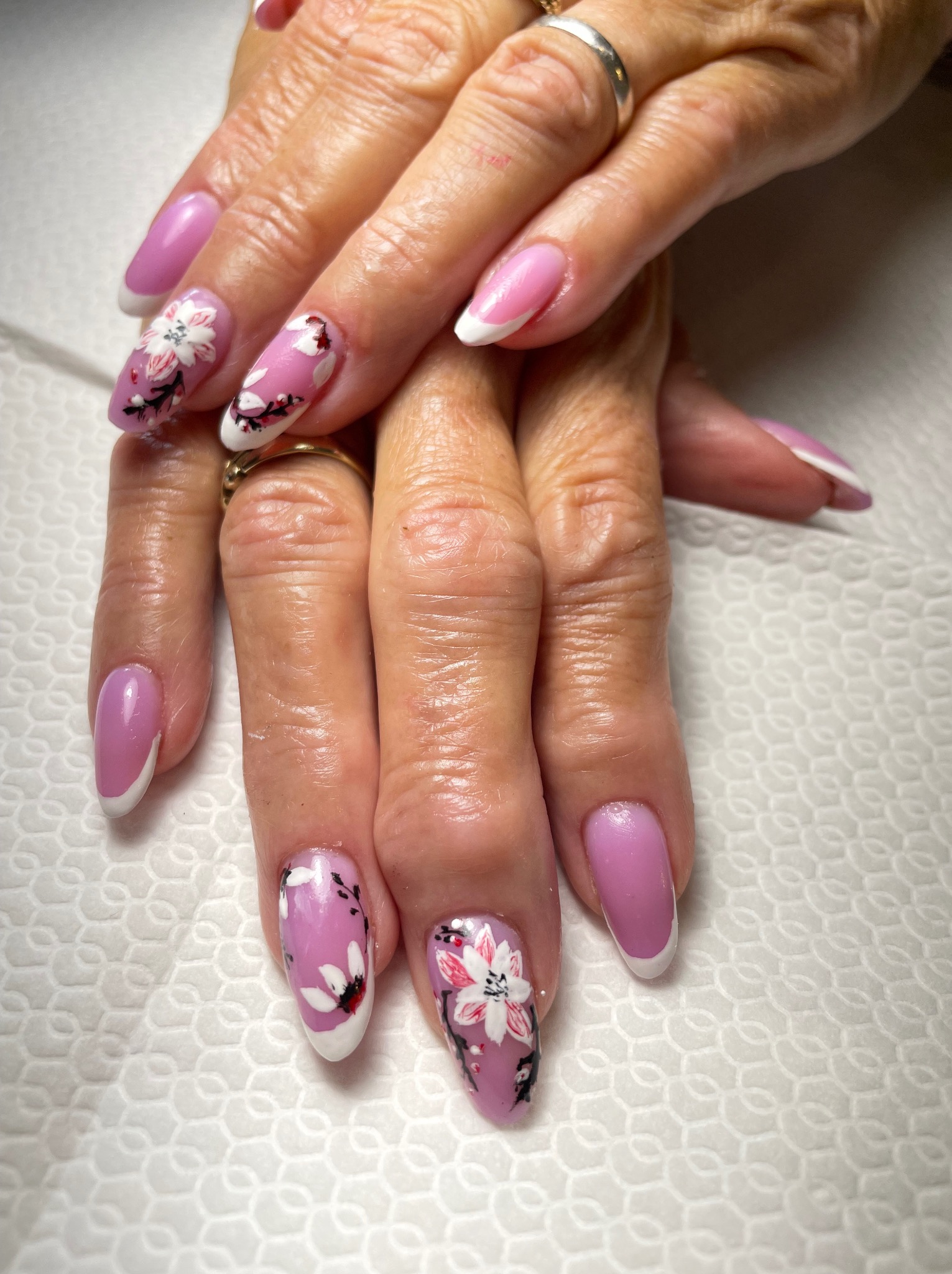 Dłonie z delikatnym, różowym manicure hybrydowym, zdobionym białymi kwiatami i francuskim wykończeniem. Zbliżenie na detale i precyzję wykonania.