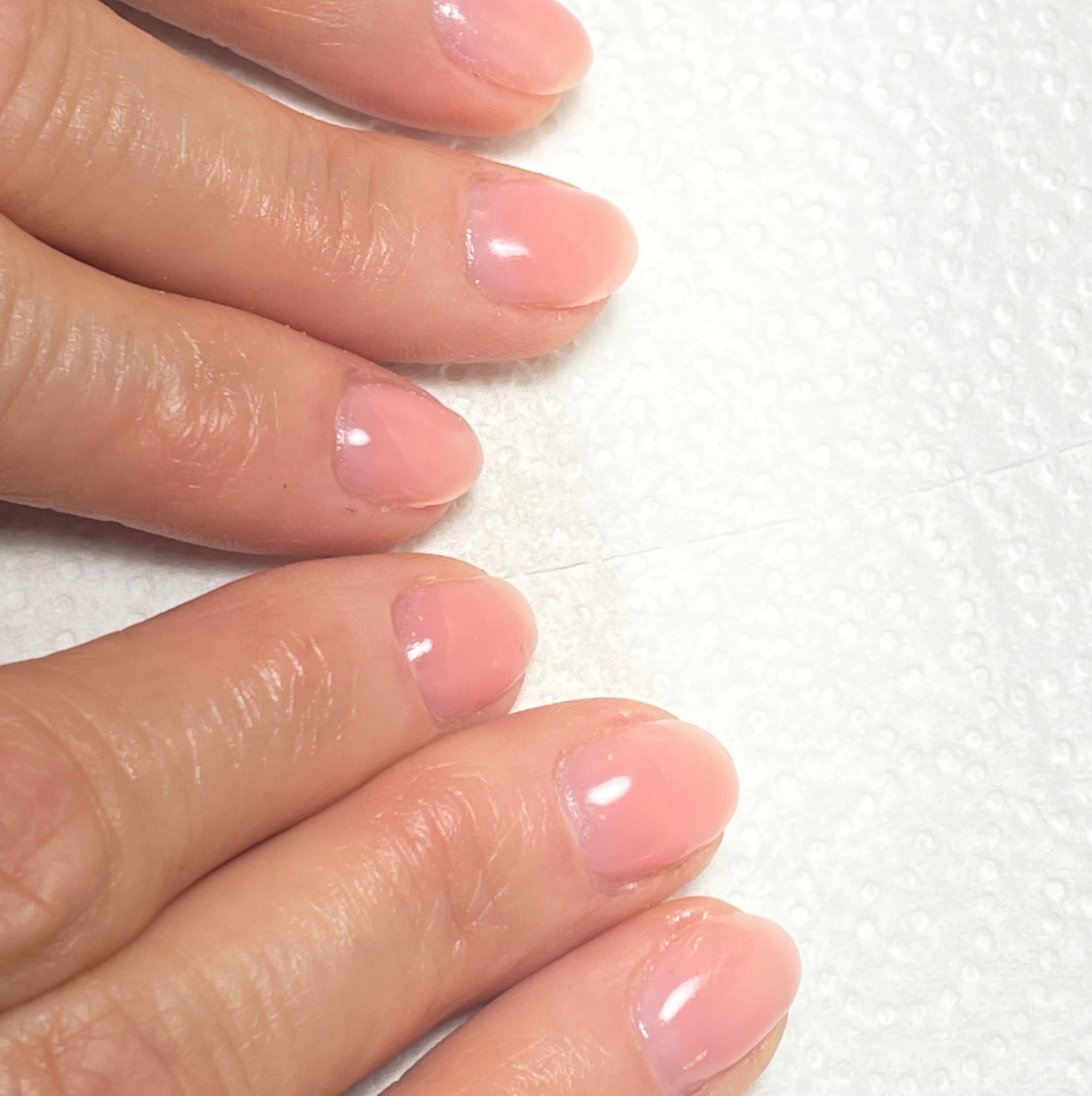 Delikatny, różowy manicure hybrydowy na naturalnych paznokciach, widoczne skórki i odbicia światła na lakierze. Tło stanowi biała, teksturowana powierzchnia.