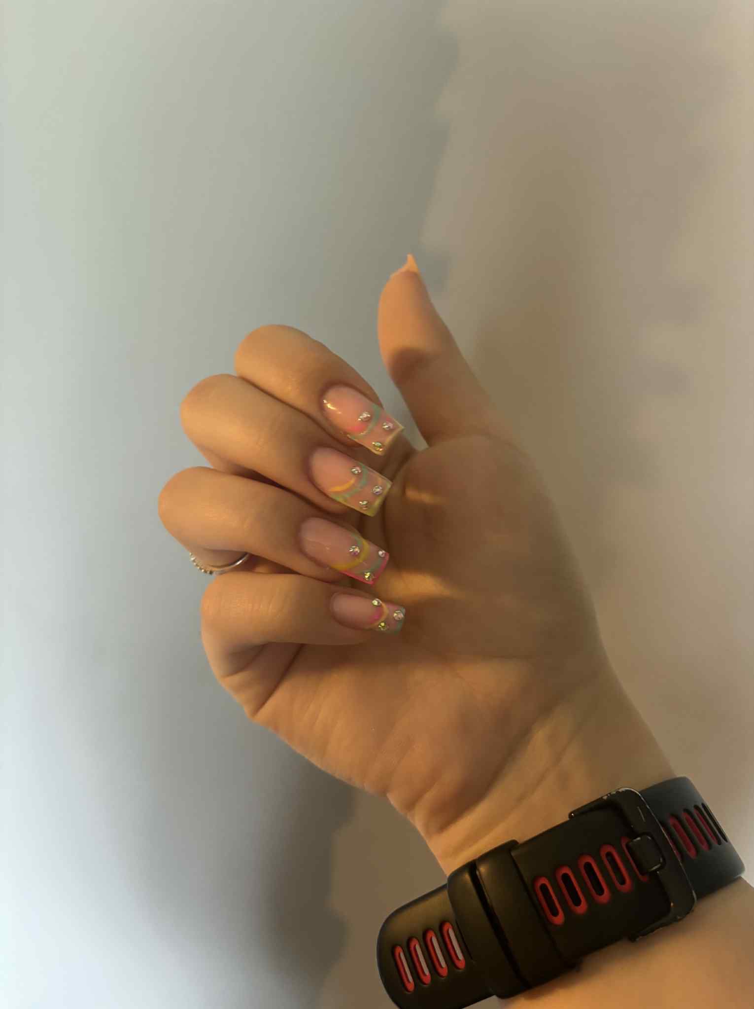 Dłoń z kwadratowymi paznokciami w pastelowych kolorach i z błyszczącymi zdobieniami na tle jasnej ściany. Widoczny zegarek na nadgarstku.