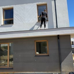 JUST PERFECT Wioleta Jałmużna - Kobieta w czarnym kombinezonie myje okno na wysokości na tle białego domu z drewnianymi ramami okiennymi i błękitnego nieba.